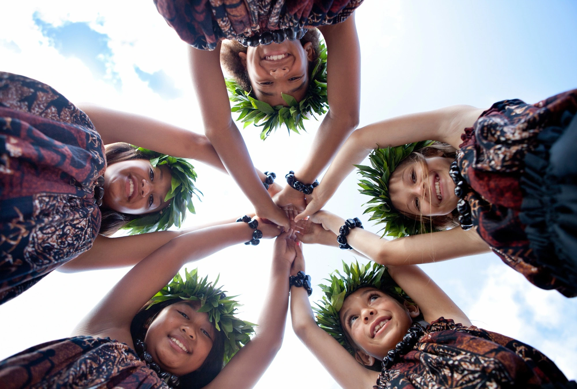 Keiki wearing haku leis join hands in a circle under a blue sky.